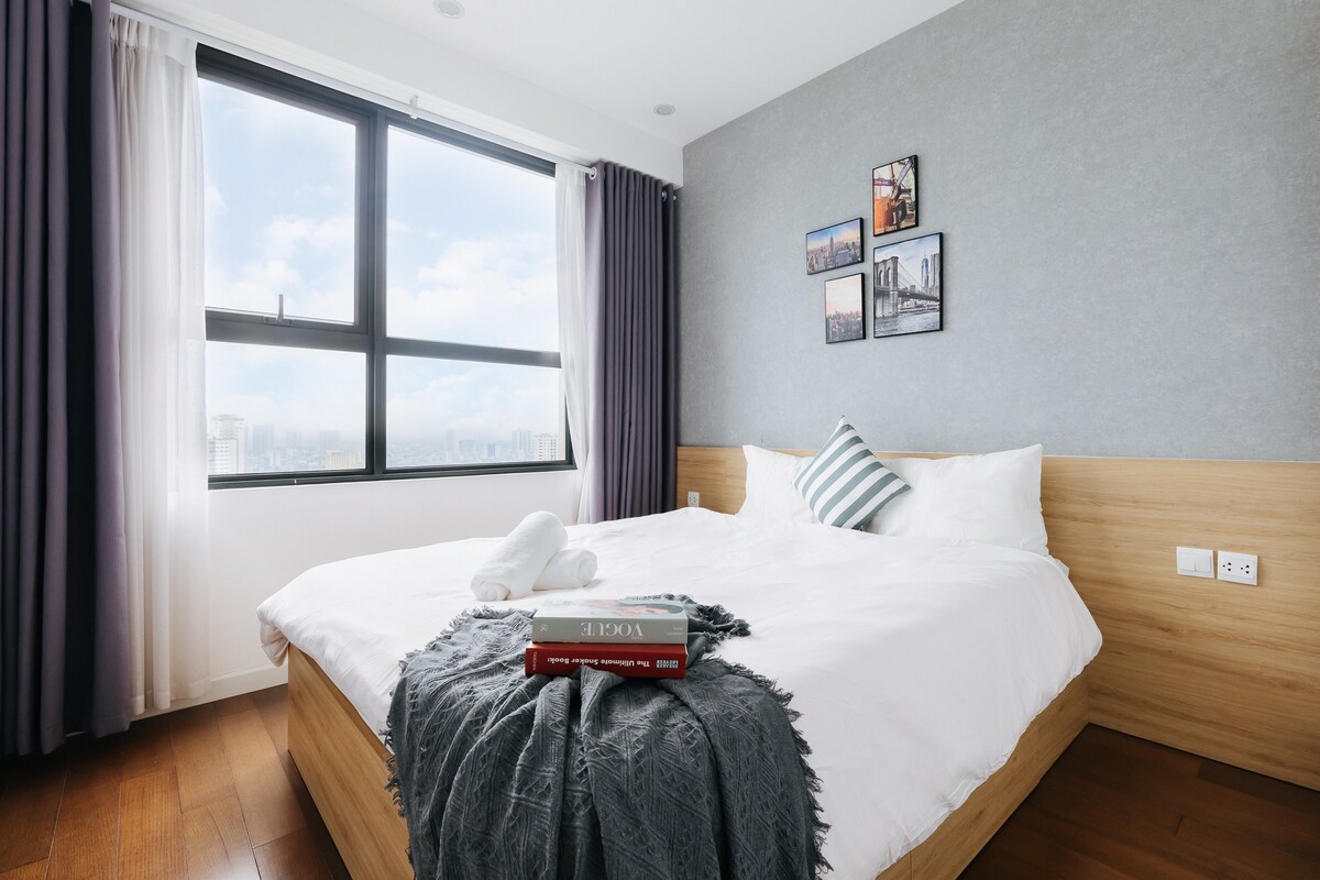A comfortable bedroom features a spacious bed with a light-colored duvet and decorative cushions. Wall art depicting cityscapes adorns the walls, and natural light filters through a large window, framed by curtains. A neatly arranged set of towels and a book is placed on the bed.