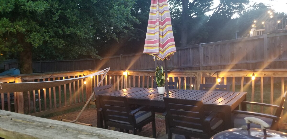 An outdoor deck area is presented, featuring a large wooden table surrounded by chairs. A colorful umbrella stands in the center, providing shade. Soft string lights are illuminated along the railing, contributing to a relaxed ambiance. A hammock is visible to the side, nestled between the trees.