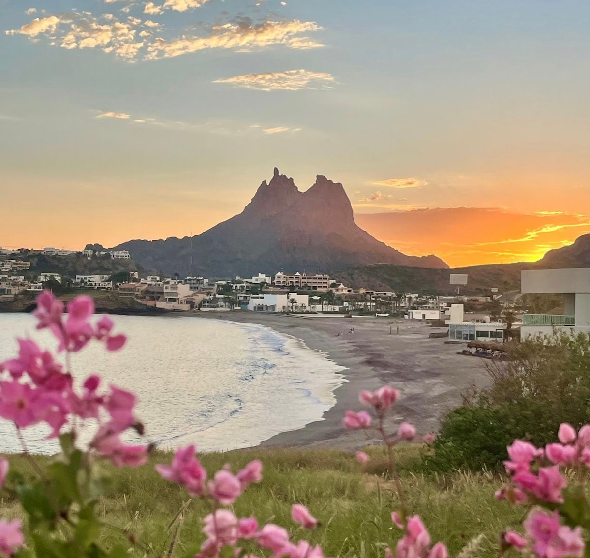 A scenic view of a beach at sunset is framed by vibrant pink flowers in the foreground. The silhouette of a mountainous landscape rises in the distance, while golden and orange hues fill the sky, reflecting on the gentle waves of the water.