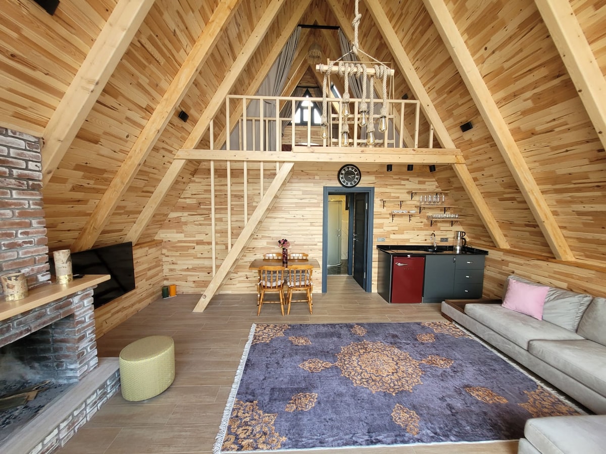 The interior features high wooden ceilings and a warm palette, with a cozy living area. A plush sofa sits on a vibrant area rug, and a wooden dining table is positioned nearby. A compact kitchen area is visible, showcasing modern appliances.