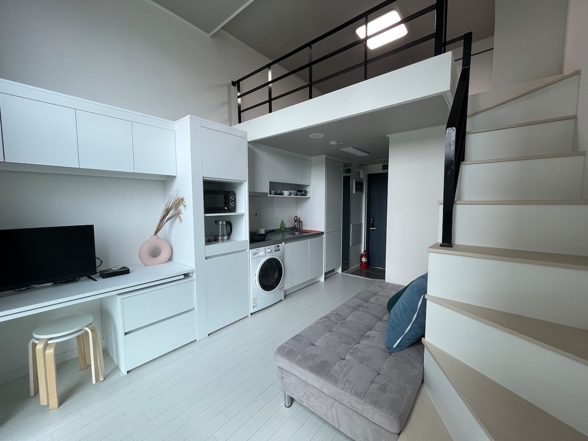An open-plan living space is featured, showcasing modern furnishings. A cozy gray sofa is positioned near a sleek kitchen area equipped with a stove and microwave. A small dining table and light-colored wood stairs leading to an upper level are visible, alongside a television on a minimalist console.
