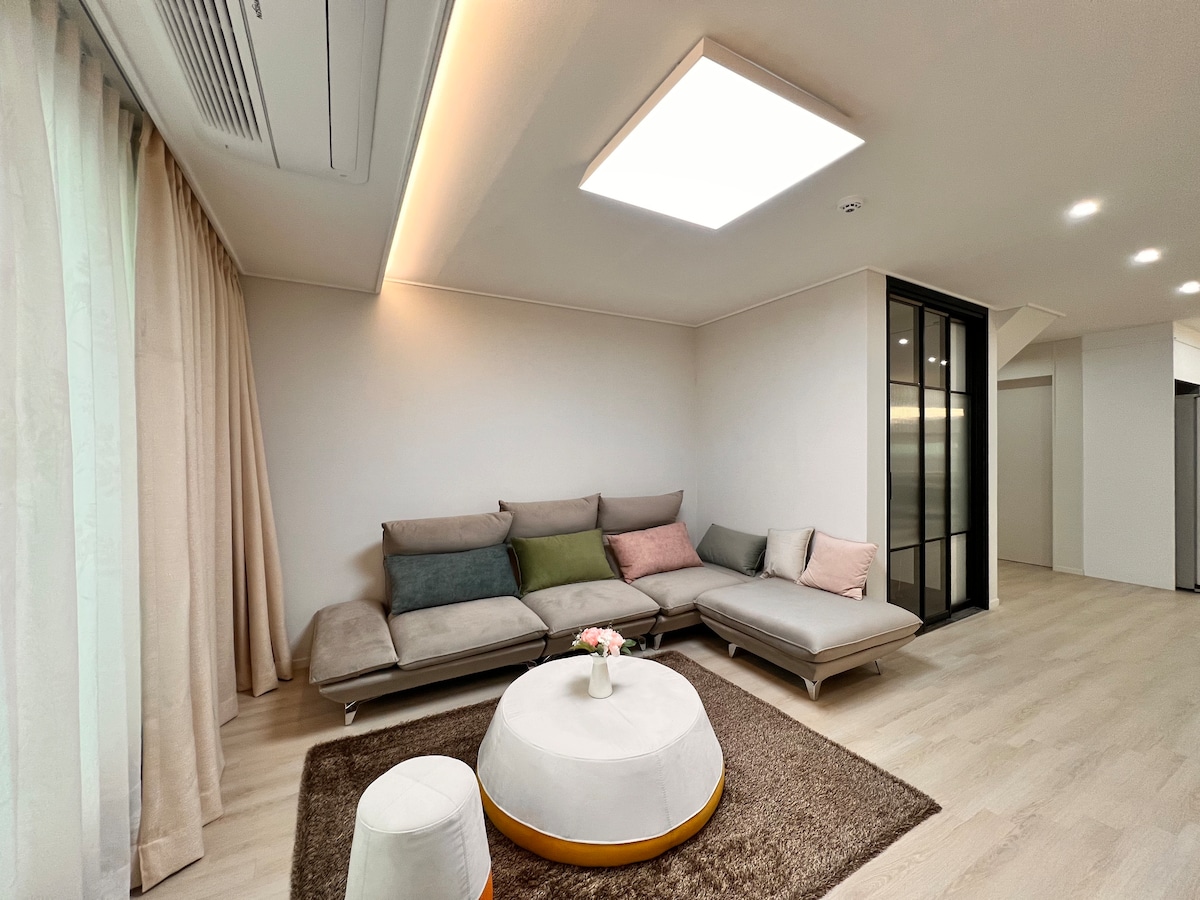 A modern living area is presented, featuring a sectional sofa with varying cushion colors, positioned around a circular coffee table. Soft natural light filters through large windows, and light-colored wood flooring enhances the airy feel of the space.