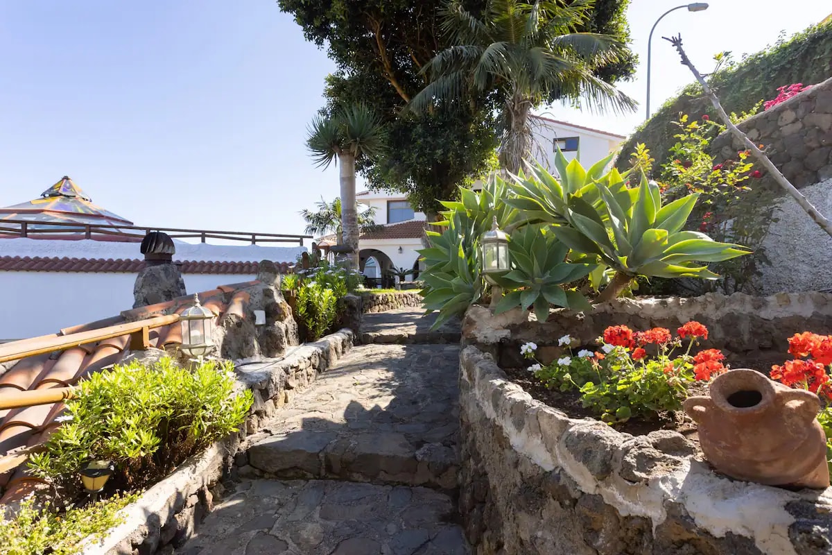 A stone pathway leads through a lush garden, flanked by vibrant flower beds featuring red blooms. Succulent plants and decorative elements, such as a clay pot, are integrated into the landscape. The area is shaded by tall trees, creating a serene atmosphere.