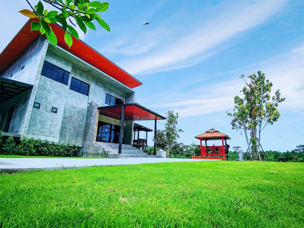 A modern building with a sleek concrete exterior features large windows and an inviting covered porch. Lush green grass surrounds the property, and a traditional gazebo is visible nearby, providing a serene outdoor space complemented by trees and open sky.