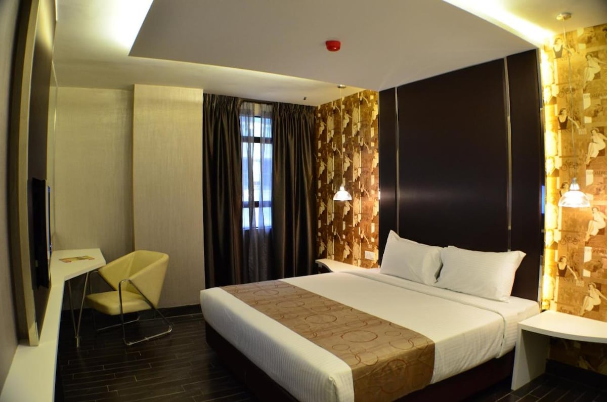A cozy room features a spacious bed with crisp white linens, complemented by decorative wall accents in warm tones. Natural light filters through the window, which is adorned with curtains. A small desk and chair are positioned near the wall, and a wall-mounted TV is visible.