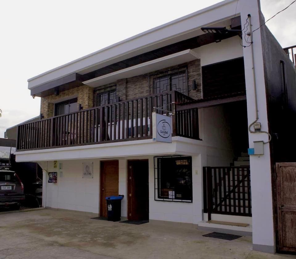 The exterior view of a two-story tourist inn features a modern design with a balcony above the main entrance. A wooden staircase leads to the upper level, while secure parking is visible in front of the building. The entrance is marked by a clear signage.