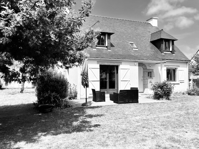 The exterior of the house is shown, with a lush green garden occupying the foreground. A patio area features dark outdoor seating, and large windows provide a view into the interior. The structure has a sloped roof and is surrounded by trees, contributing to a serene environment.