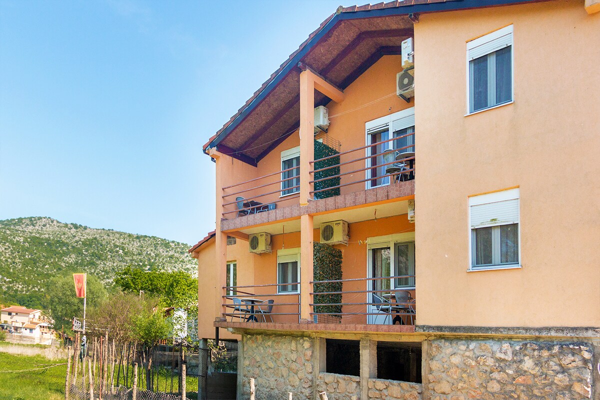 A three-story building is depicted, featuring a warm exterior with peach-colored walls and a stone ground level. Two balconies with chairs overlook the landscape, and air conditioning units are visible. Green hills rise in the background under a clear blue sky.