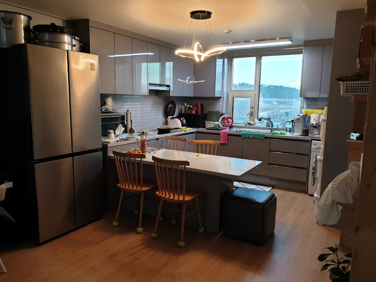 A modern kitchen features sleek cabinetry and a large island with seating. A silver refrigerator and various appliances are visible. Natural light streams through a window above the sink, and utensils and kitchenware are neatly arranged on the counter.