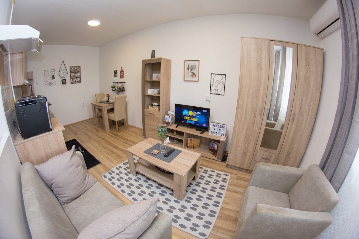 A modern living area features a light-toned wooden coffee table placed on a patterned rug. Two comfortable armchairs and a sofa are arranged around the table. A flat-screen television is situated on a console. Natural light floods the space through a nearby window.