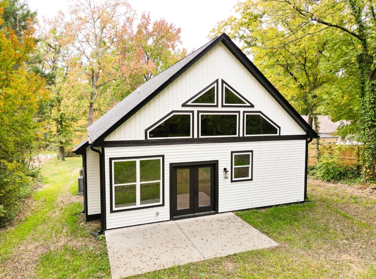 The exterior of the house is displayed, showcasing a modern design with a combination of white siding and black trim. Large, angled windows dominate the front facade, allowing natural light to fill the interior. A concrete patio leads into the home, surrounded by greenery.