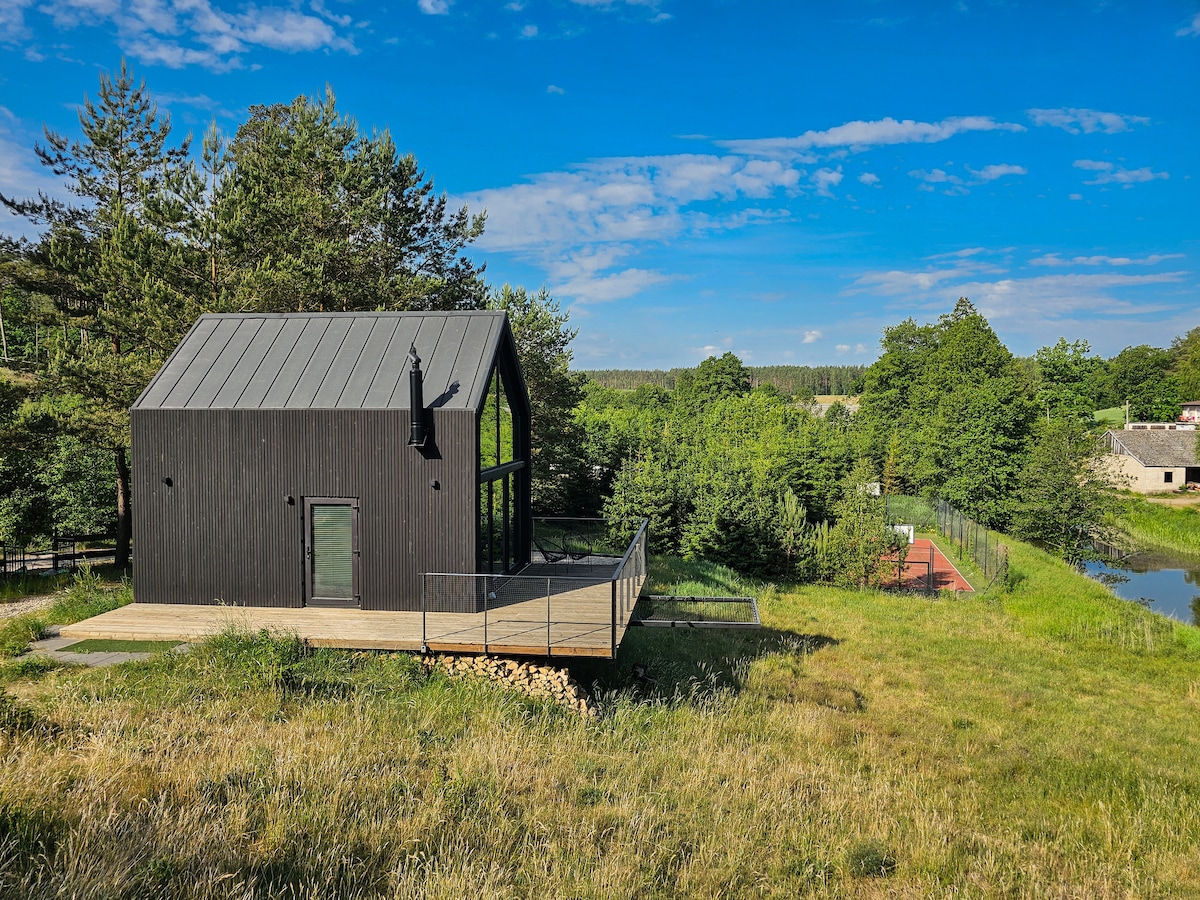 A modern cottage features a sleek black exterior and large glass windows. Surrounding greenery includes tall trees and grassy areas. An expansive deck extends from the cottage, offering views of the serene landscape and nearby water.