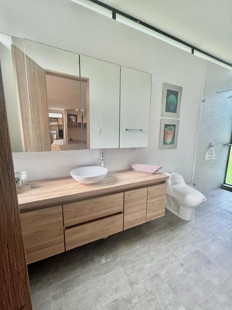A modern bathroom is featured with a wooden vanity, complemented by a round basin. A large mirror enhances the openness of the space. A white toilet is positioned nearby, and textured tiles create a contemporary look. Natural light enters through a window, illuminating the room.