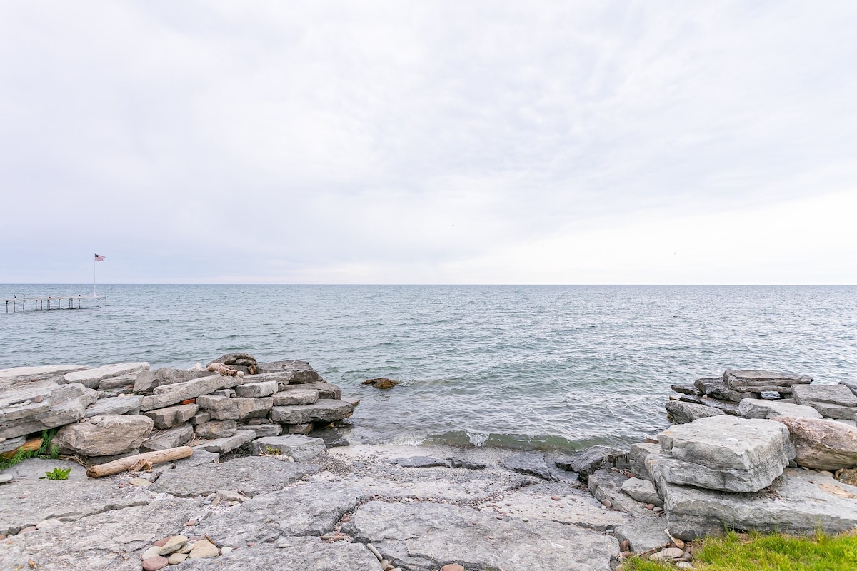 The image captures a serene shoreline with smooth stone formations leading to calm waters. The horizon features a gentle blend of sky and sea, while a flagpole stands in the background, suggesting nearby recreational facilities. The atmosphere conveys a sense of tranquility.
