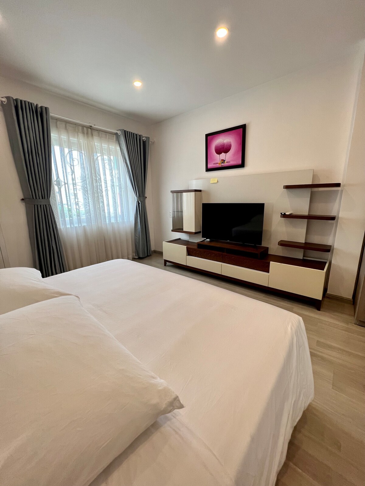 A cozy bedroom setting features a spacious bed with crisp white linens. A modern television is positioned on a stylish unit, complemented by a decorative wall artwork. Natural light enters through sheer curtains, enhancing the bright and calming atmosphere.