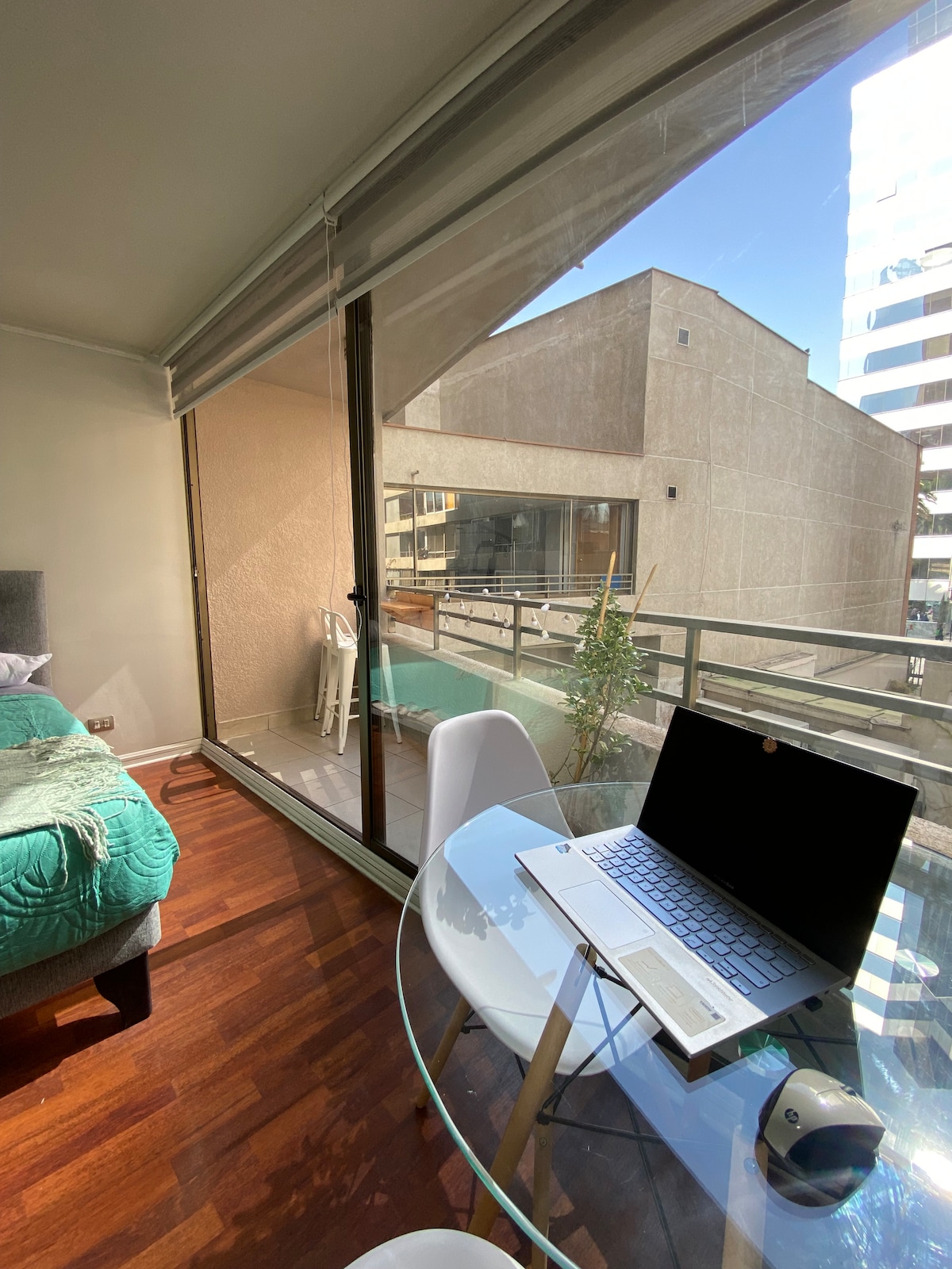 A work desk is positioned beside a large window, allowing natural light to fill the space. A laptop sits on a glass table, accompanied by a small plant. The room features a cozy bed with a green blanket, and a balcony is visible outside.