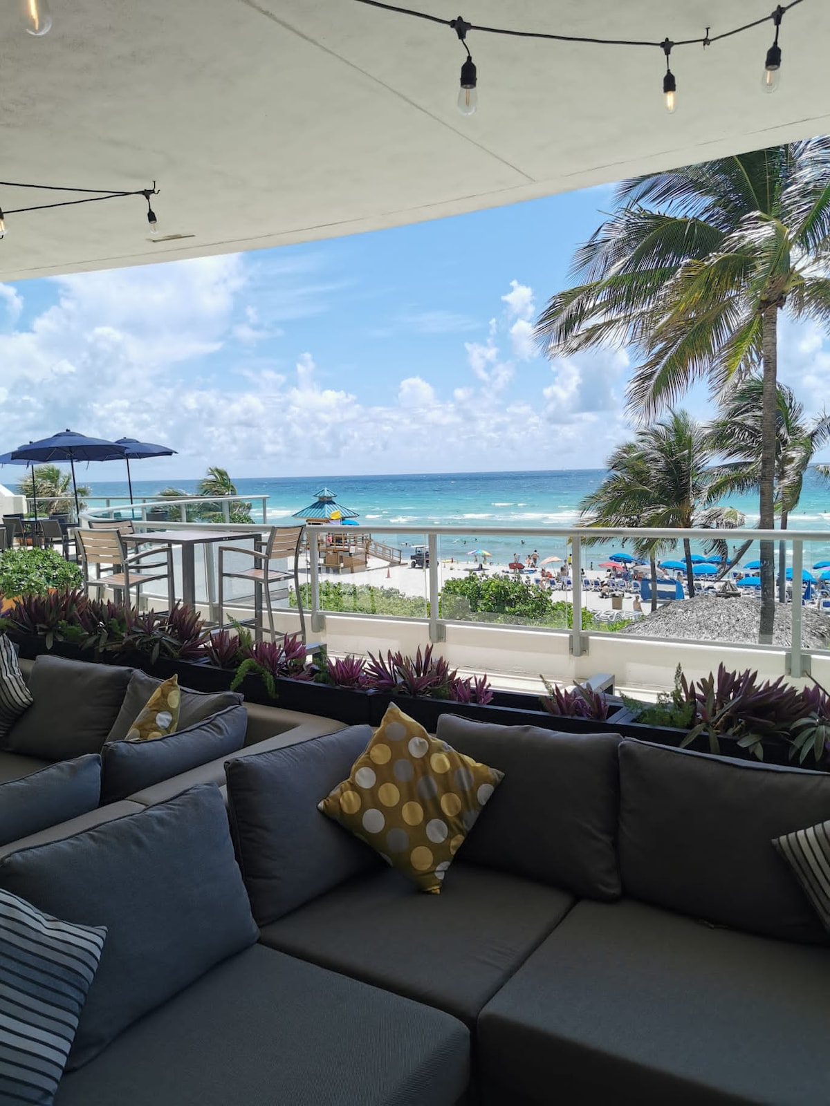 A spacious outdoor seating area is visible, featuring plush gray couches adorned with patterned pillows. The backdrop reveals a sweeping view of the turquoise ocean and beach, framed by swaying palm trees and a clear blue sky.