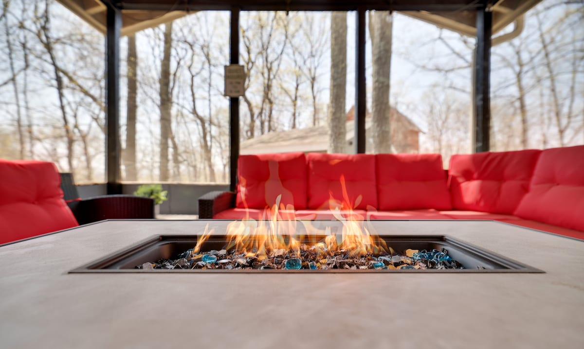 A gas fire pit is centered on a large table, with flames flickering above colorful glass stones. Surrounding seating features bright red cushions, providing a comfortable area for relaxation. The backdrop reveals trees with budding leaves, suggesting a serene outdoor setting.