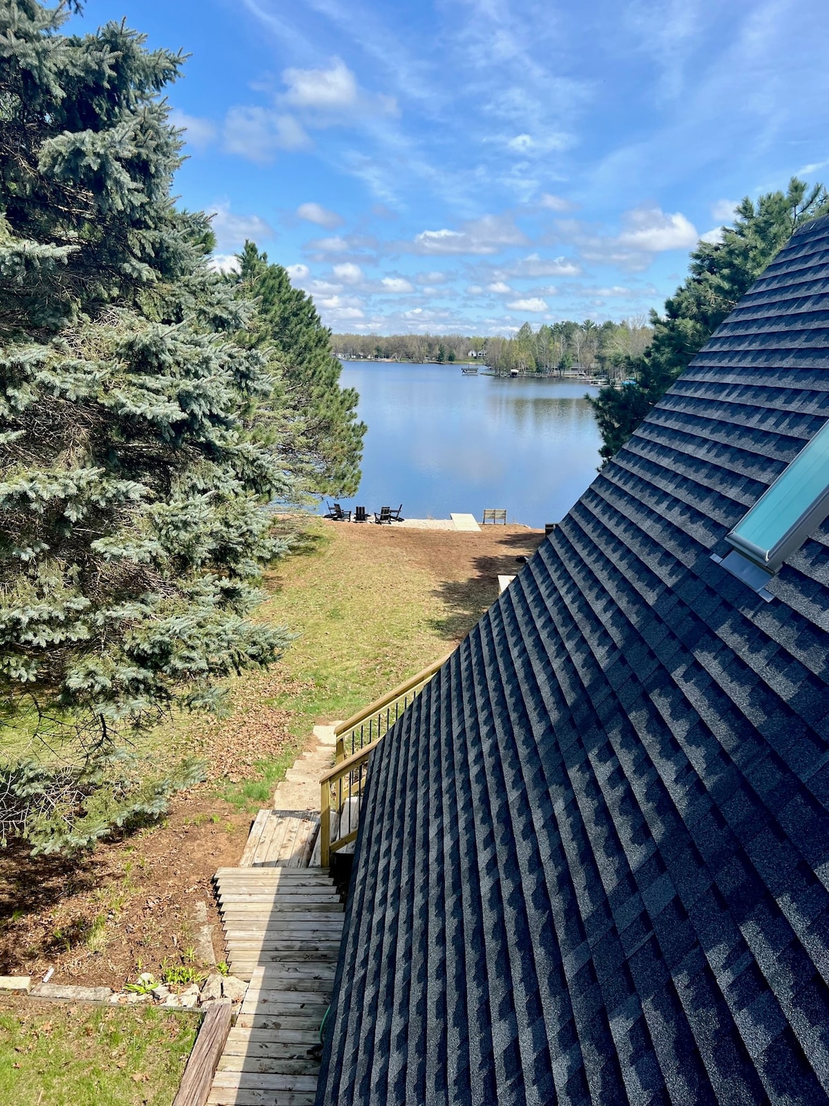 A serene view of the lake is framed by tall pine trees. A grassy yard leads down to the water's edge, where seating is arranged for lakeside enjoyment. The A-frame shape of the house provides a unique angle, showcasing the peaceful natural surroundings and clear blue sky.