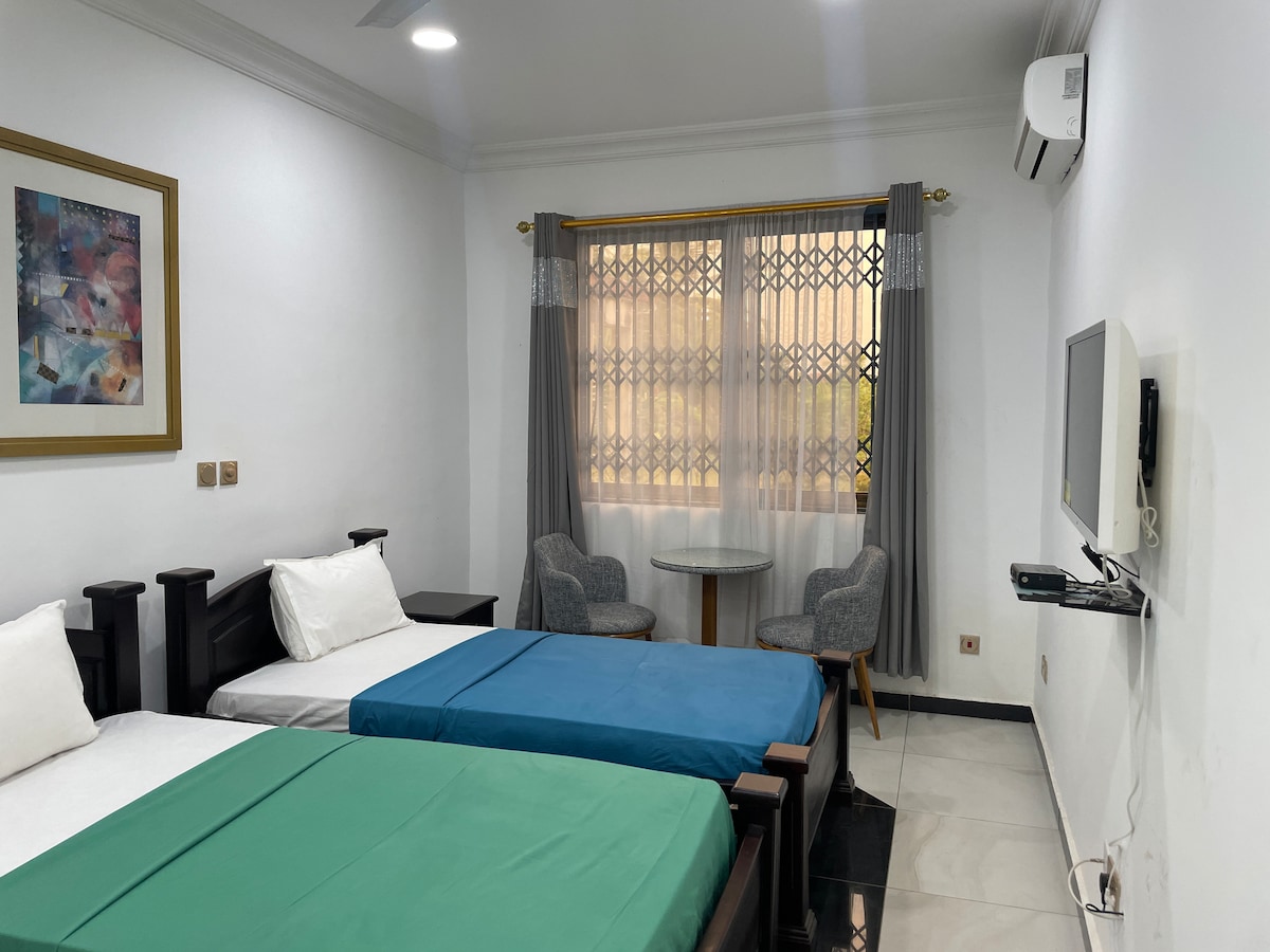 A bright room features two spacious beds, one covered with a green sheet and the other with a white sheet. A small round table with two chairs is positioned near a window adorned with sheer and solid curtains. A wall-mounted television is visible opposite the beds.