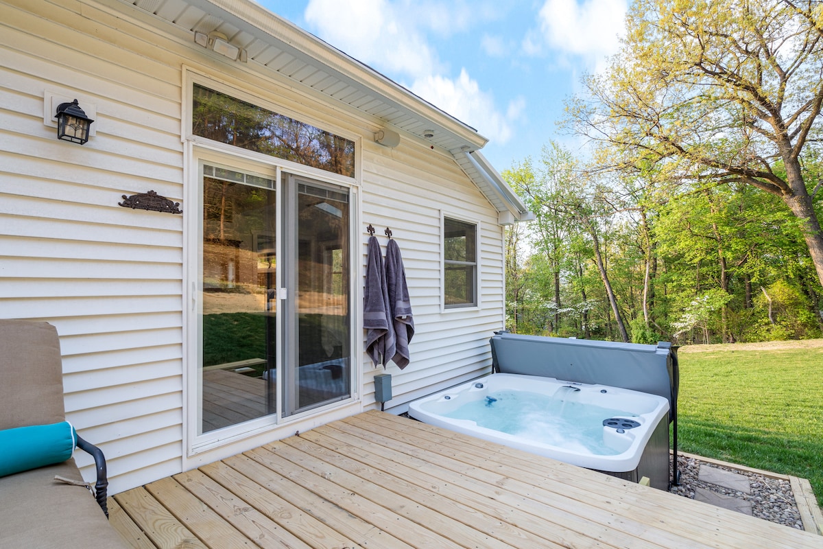 A private deck is shown featuring a jetted hot tub surrounded by wooden decking. Soft towels hang on hooks nearby, with a spacious lawn visible in the background, bordered by trees that provide natural shade.