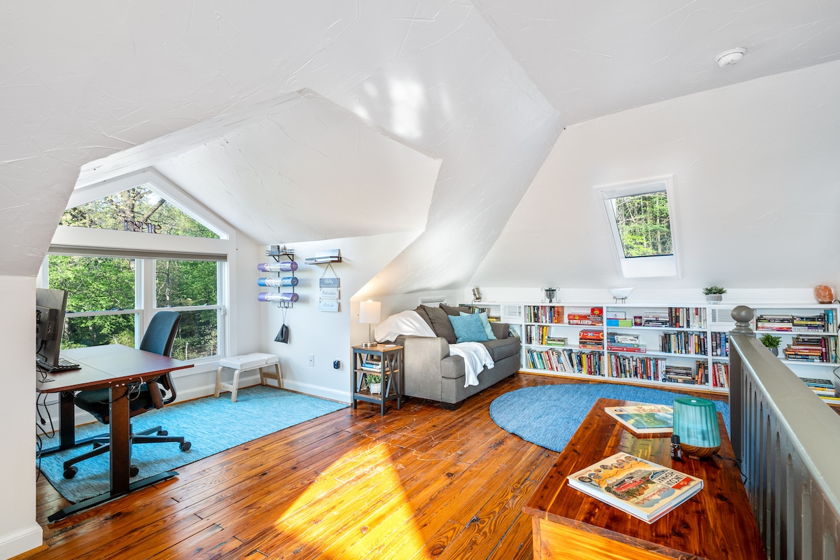 A multifunctional room displays a modern aesthetic, featuring natural wood floors and sloped ceilings. A cozy sitting area with a pullout sofa and plush rug is arranged near a bookshelf filled with various titles. A desk and chair are positioned for work, complemented by skylights that allow sunlight in.