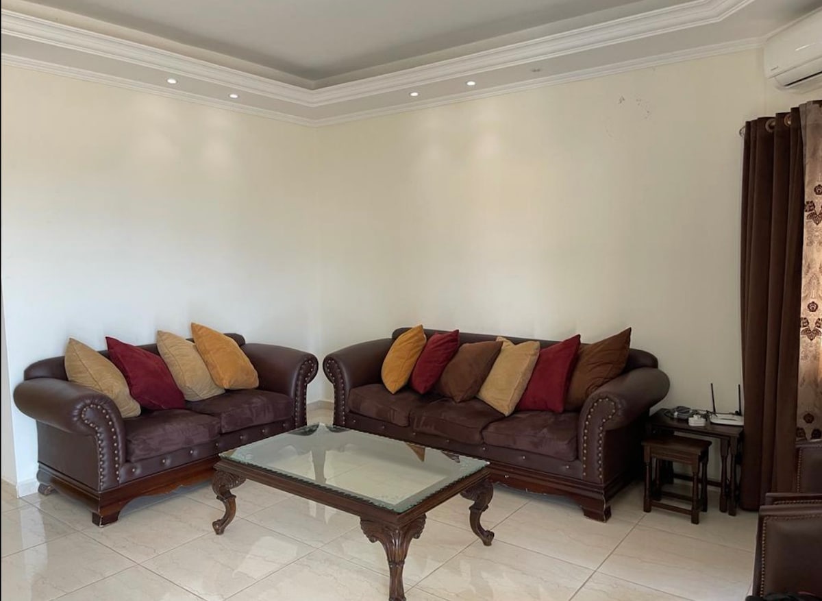 A cozy living area is furnished with two dark leather sofas adorned with an assortment of colorful cushions. A glass coffee table sits in the center, reflecting room light. Soft lighting from the ceiling enhances the inviting atmosphere, while dark curtains frame a window.