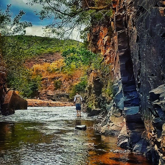 A tranquil river scene depicts a person walking along the water's edge, surrounded by tall rocky cliffs. Lush greenery frames the river, highlighting the natural beauty of the area. The clear water reflects the surrounding landscape under a partly cloudy sky.