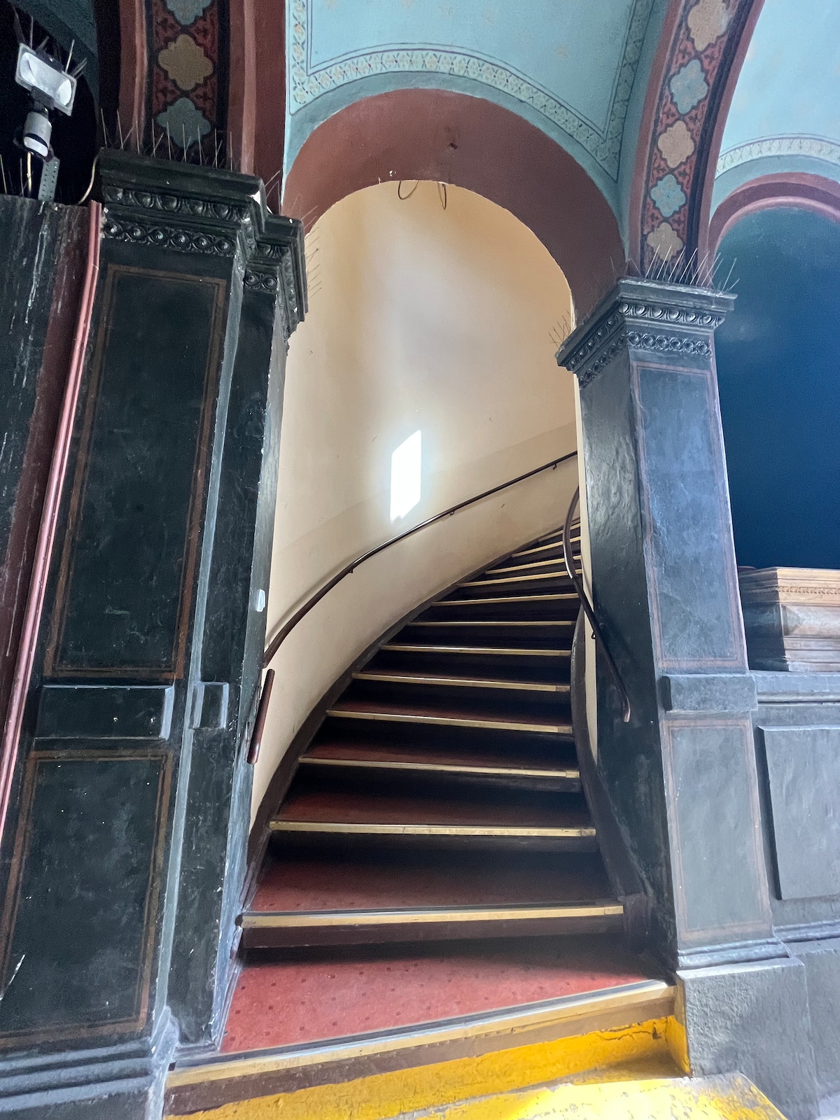A winding staircase features wooden steps leading to an upper level. The staircase is flanked by ornate architectural columns, and a small window at the top allows light to filter through, illuminating the space while highlighting the details of the structure.