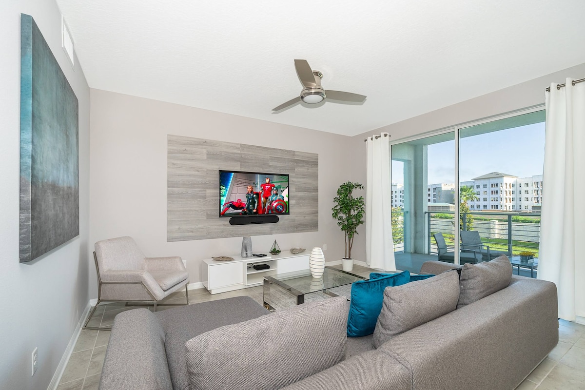 A spacious living room is shown, featuring a large, sectional sofa with blue accent pillows. A flat-screen TV is mounted on a textured wall, and a light-colored chair is positioned nearby. Natural light enters through sliding glass doors that lead to a private balcony.