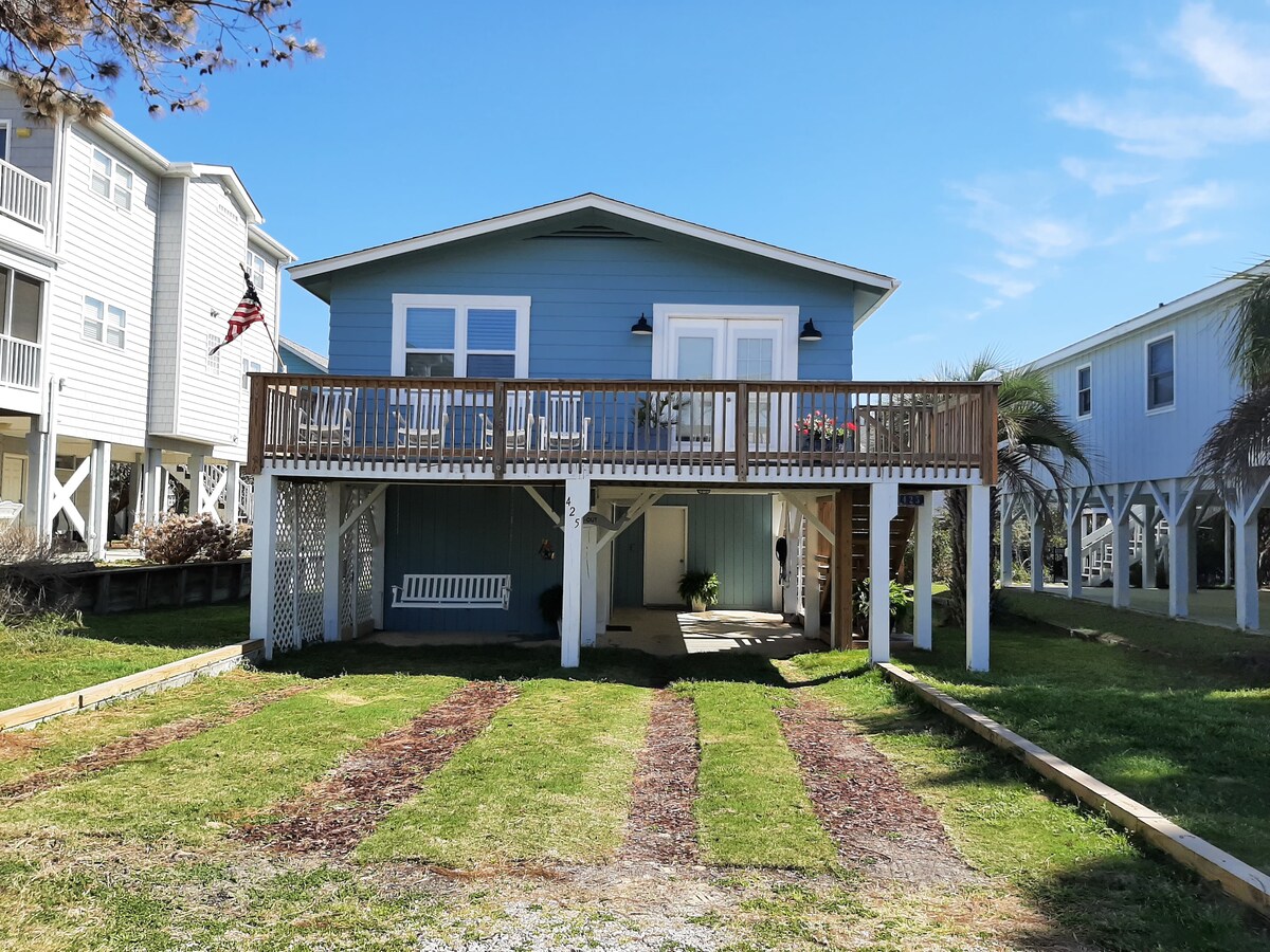 A charming two-story cottage features a light blue exterior and a spacious front porch adorned with potted plants. The structure is elevated on stilts, allowing for shaded parking underneath. Simple landscaping with a grassy yard provides a welcoming approach to the entrance.