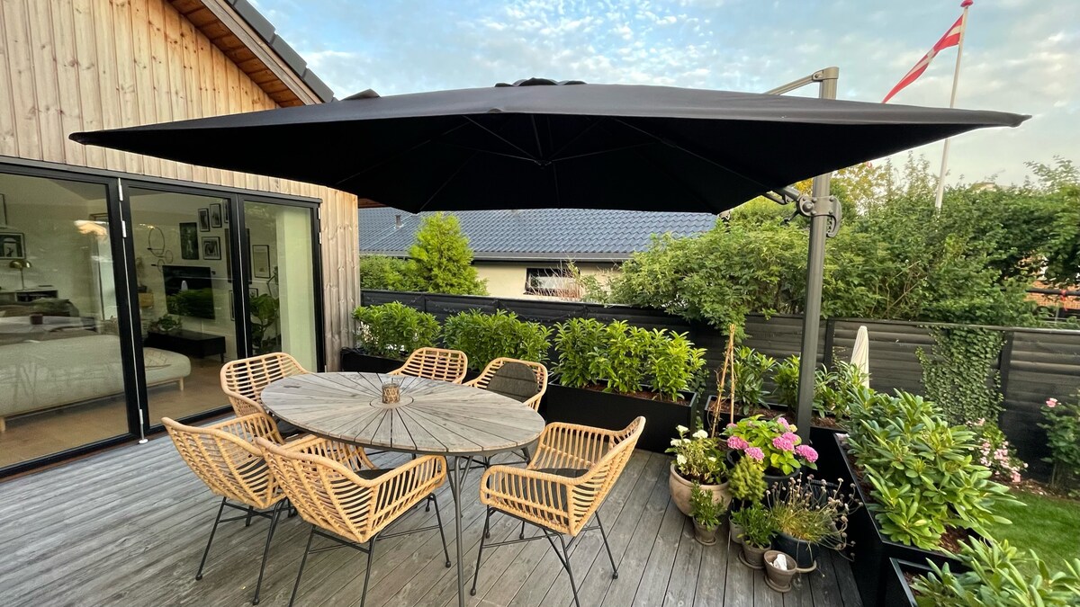 An outdoor terrace features a round wooden table surrounded by six woven chairs, shaded by a large black umbrella. The area is adorned with various plants in planters, contributing to a natural ambiance, with a wooden deck providing a smooth surface.