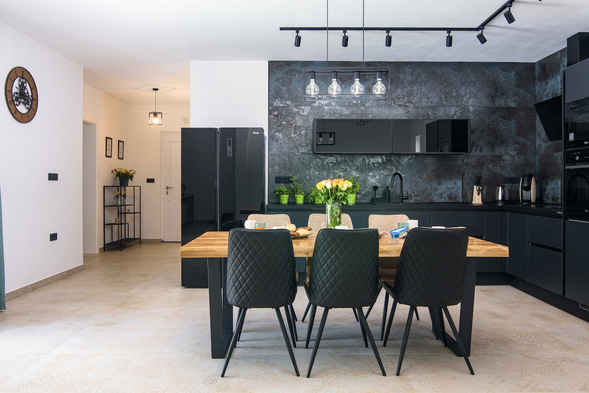 A sleek kitchen area features a dark stone accent wall behind modern cabinetry. A wooden dining table with six black chairs is centered, accompanied by a vase of fresh flowers. Light fixtures hang above, illuminating the open space that connects with a well-organized pantry.