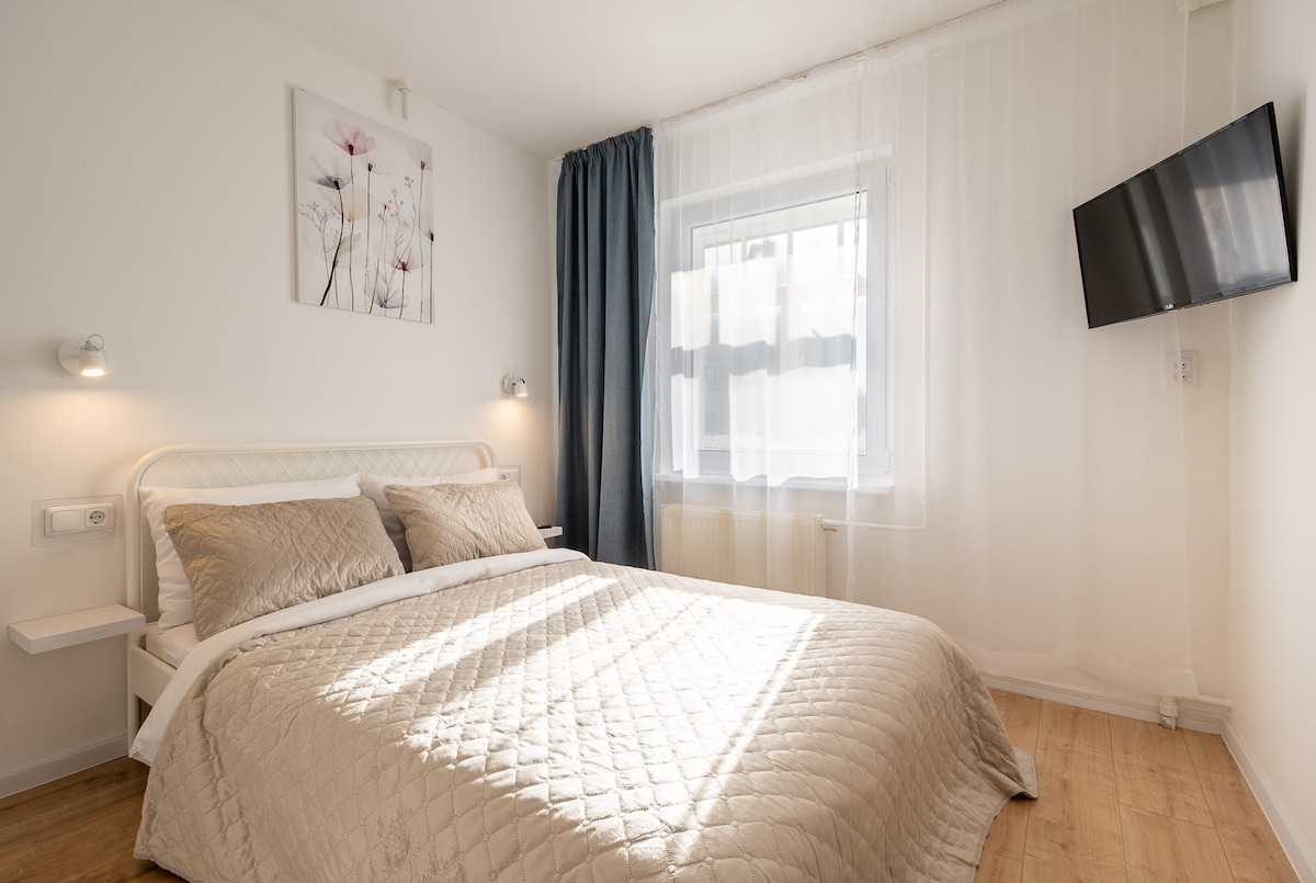 A bright bedroom is presented with a comfortable bed featuring a quilted cover and decorative pillows. Natural light filters through a window adorned with sheer curtains. A small television is mounted on the wall, and soft light is provided by wall sconces on either side of the bed.