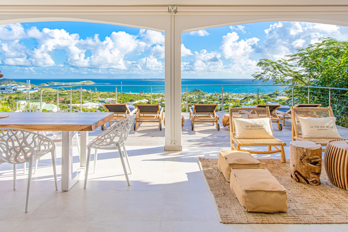 An airy terrace is showcased with views of the sea and sky. The space features a wooden dining table alongside white chairs, complemented by lounge chairs and natural decor elements. Bright light fills the area, enhancing the spacious and relaxed atmosphere.