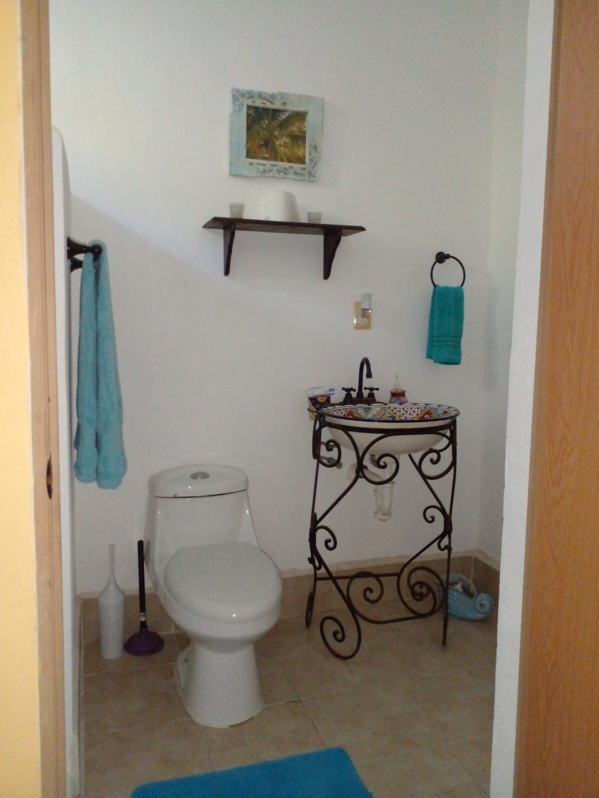 A private bathroom is presented, featuring a modern toilet and a decorative sink with an elegant metal stand. A small shelf holds toiletries, while towels hang on nearby hooks. Soft blue mats add a touch of comfort to the tiled floor.