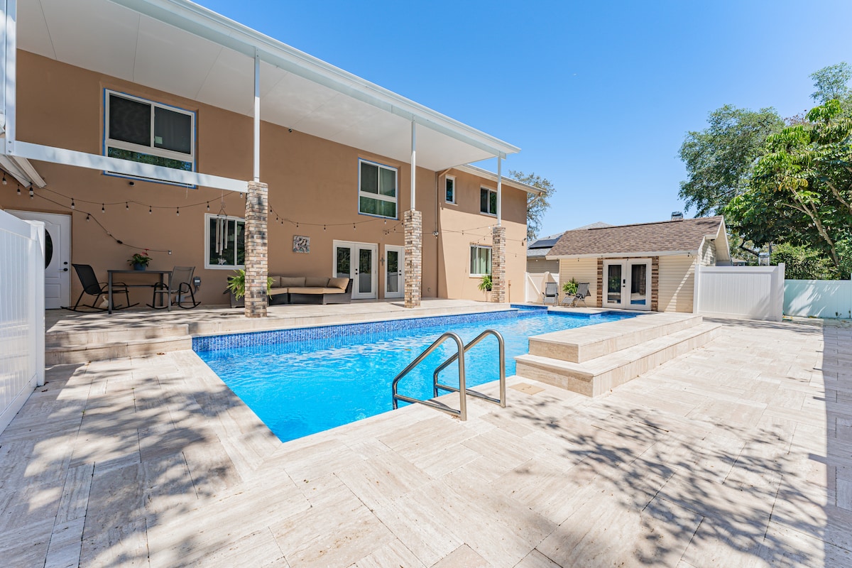 A spacious backyard features a large saltwater pool surrounded by ample deck space. The area is framed by stone pillars and is complemented by a backdrop of a two-story residence. Nearby, comfortable seating is visible, inviting relaxation in the sunny setting.