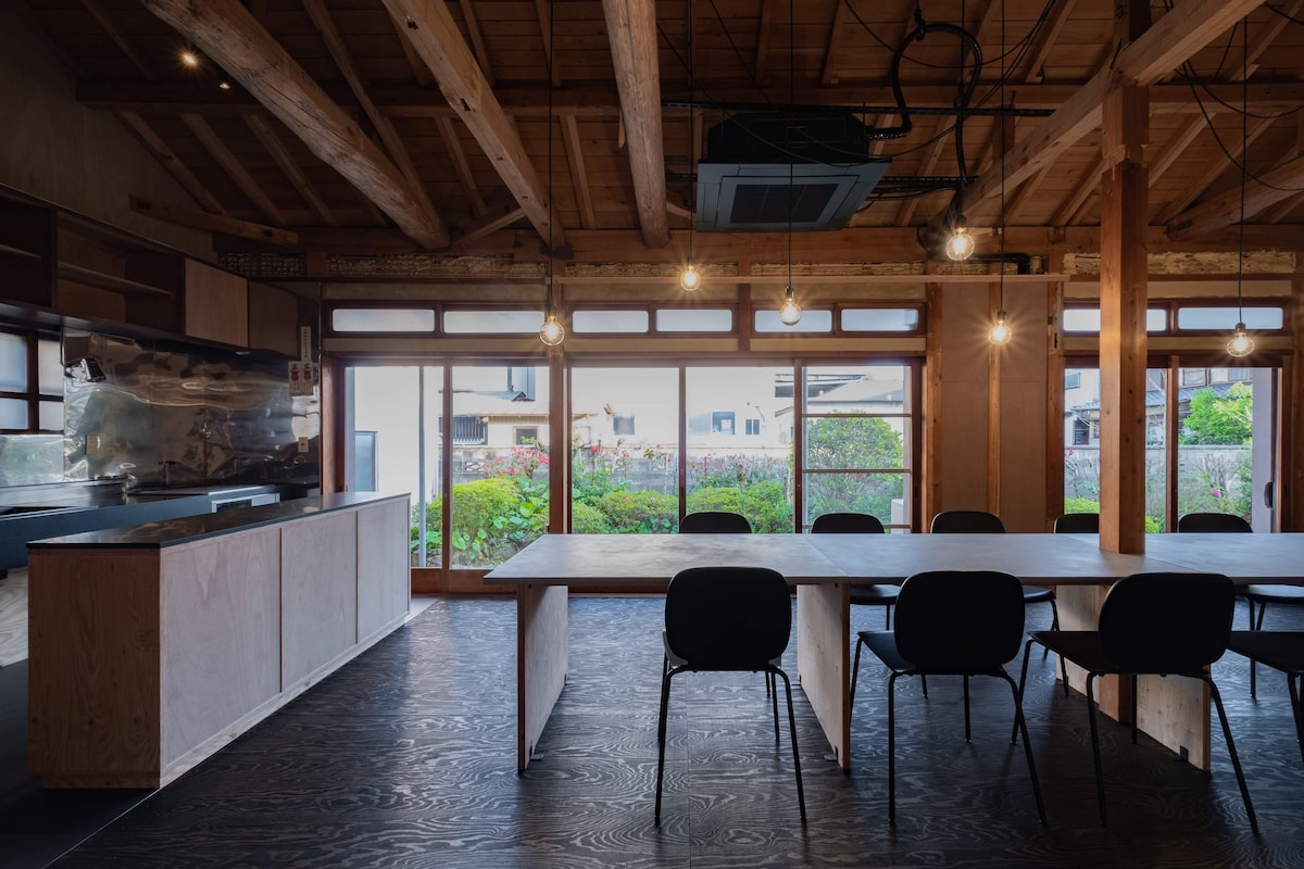 A spacious dining area features a long wooden table surrounded by black chairs. Large windows provide a view of the garden and allow natural light to fill the room. Exposed beams and pendant lighting add to the rustic charm of the renovated space.