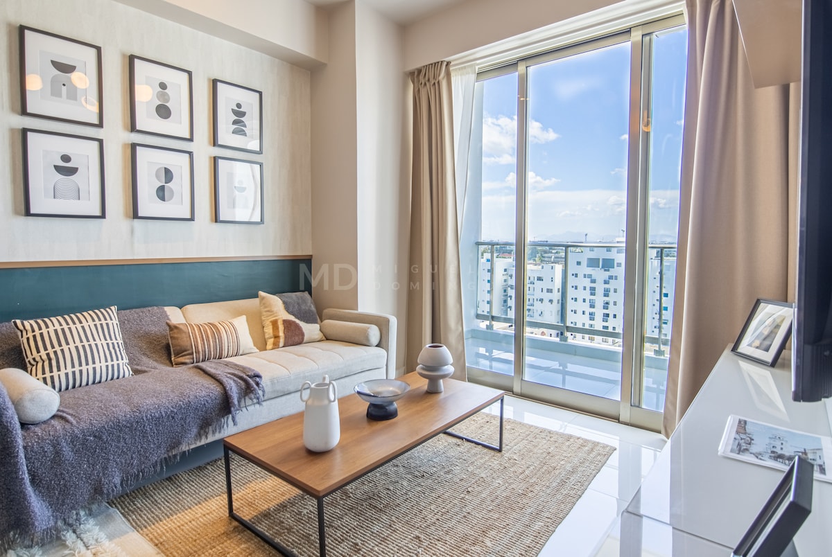 A cozy living area features a comfortable sofa adorned with throw pillows and a textured blanket. A wooden coffee table is positioned in front, displaying decorative items. Large windows invite natural light and offer a view of the city, complemented by soft curtains.