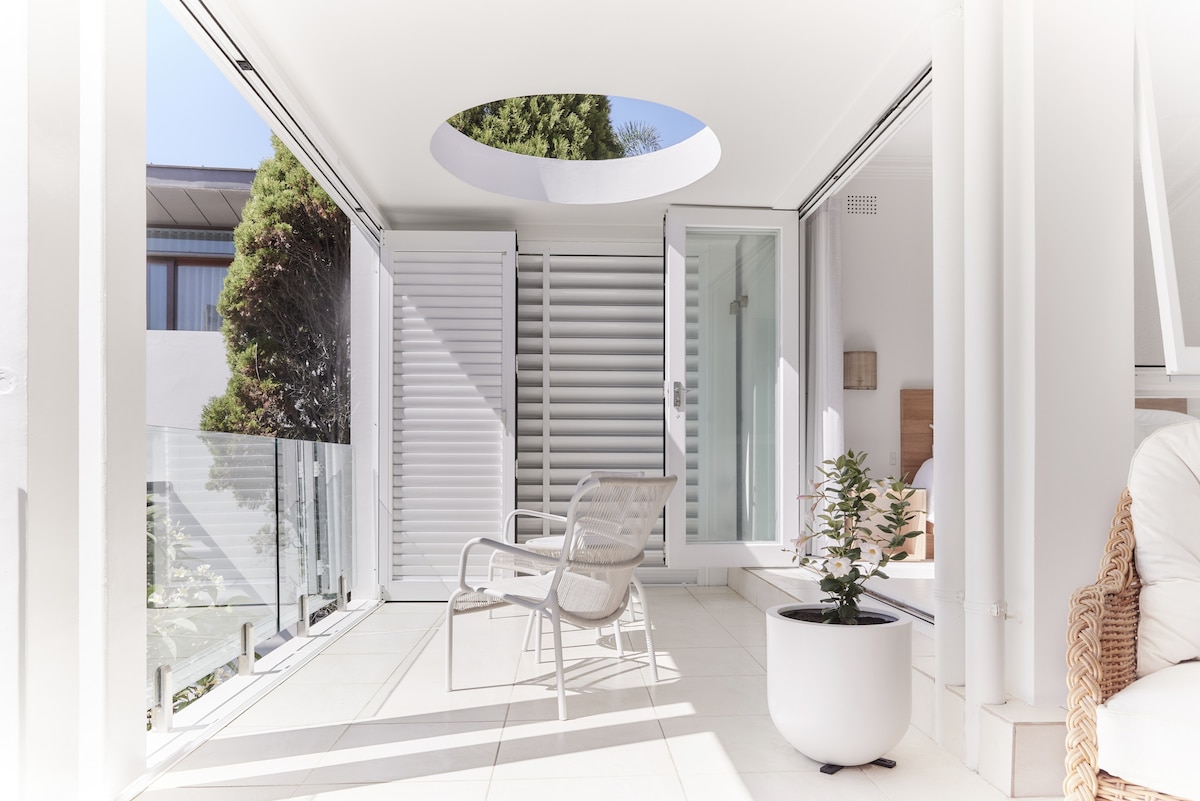 A bright and airy space is showcased, featuring a minimalistic design with large sliding glass doors that open to the outdoors. A comfortable chair and a potted plant enhance the serene atmosphere, while natural light filters through a round skylight above.
