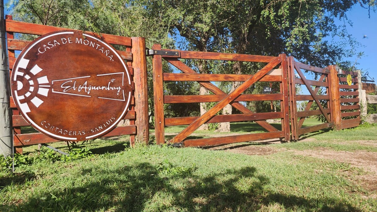 A wooden entrance gate features a branded sign for 'Casa de Montañal' and clearly displays the name 'El Efluvio.' The lush green grass surrounding the gate indicates a serene outdoor setting, with trees visible in the background.