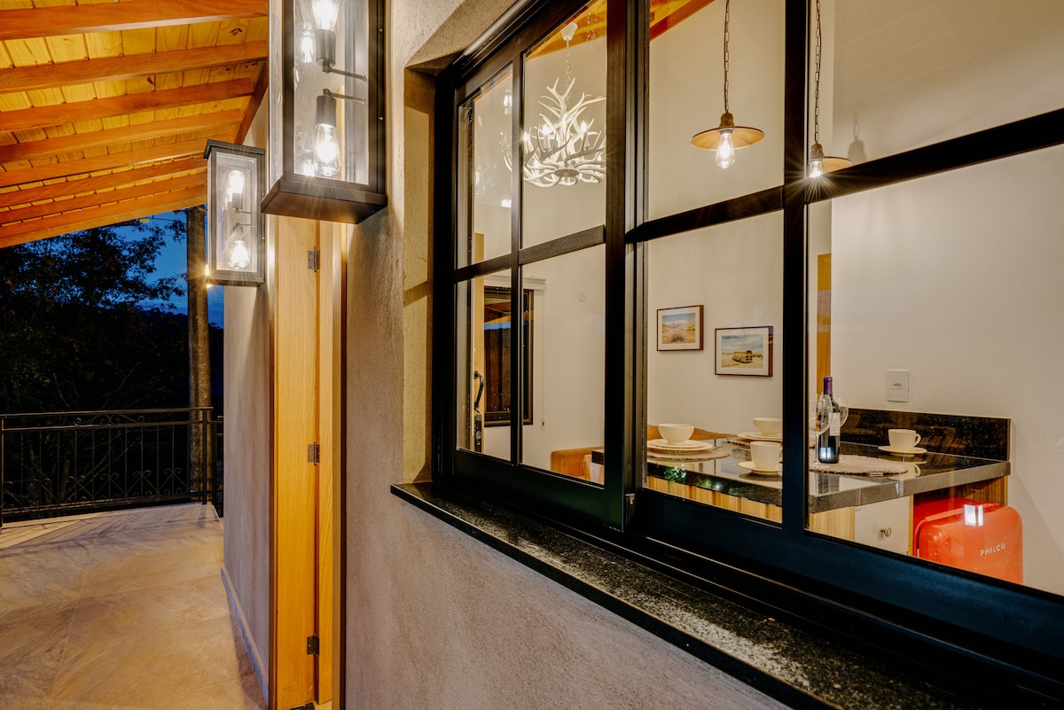 The exterior of the cabin is softly illuminated as night approaches, revealing large windows that provide a glimpse into the dining area. A cozy indoor setting is visible, complemented by warm lighting fixtures and rustic decor, creating an inviting atmosphere.