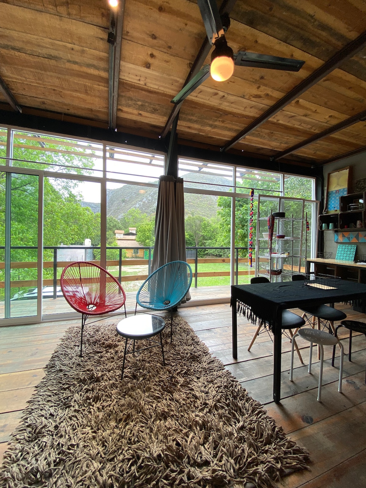 A cozy seating area is highlighted by two colorful chairs positioned on a soft rug, with a view of greenery and mountains visible through large windows. A dining table with chairs is present, complemented by warm, wooden accents throughout the space.
