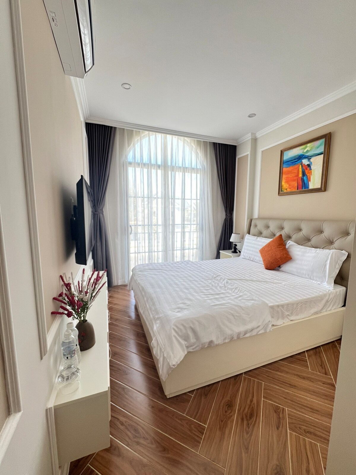A well-appointed bedroom features a spacious bed with crisp linens and an orange accent pillow. Natural light filters through large, arched windows, complemented by elegant drapes. A small artwork adorns the wall above the bed, and a television is mounted nearby.