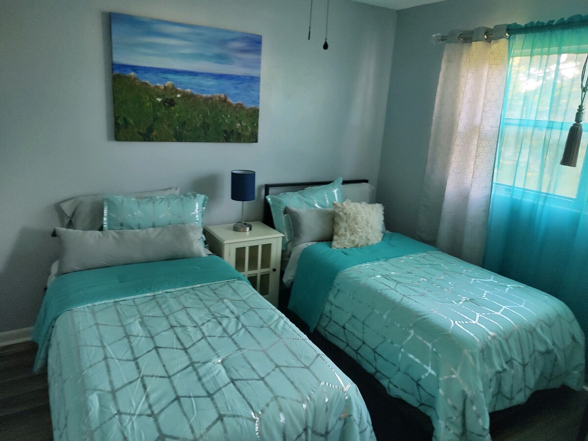 The sleeping area features two twin beds covered with light teal quilts. A framed landscape painting adorns the wall above the beds. A bedside table with a blue lamp is positioned between the beds. Natural light filters through sheer curtains on the window.