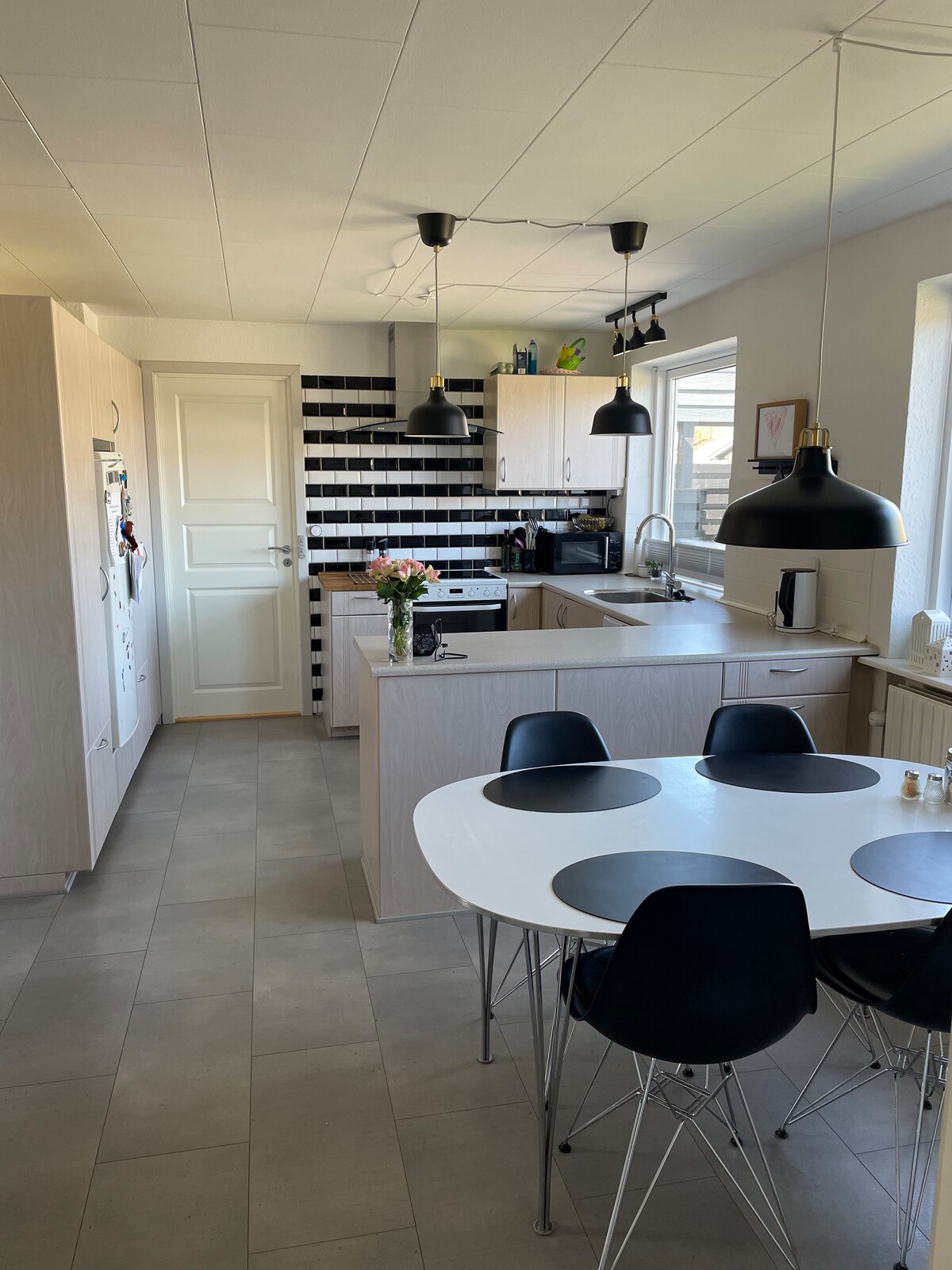 A modern kitchen features light wood cabinetry with a sleek countertop. Black and white tiles create a stylish backsplash. The space is illuminated by pendant lights, and a round dining table with black chairs offers seating for several. Natural light floods in through a window.