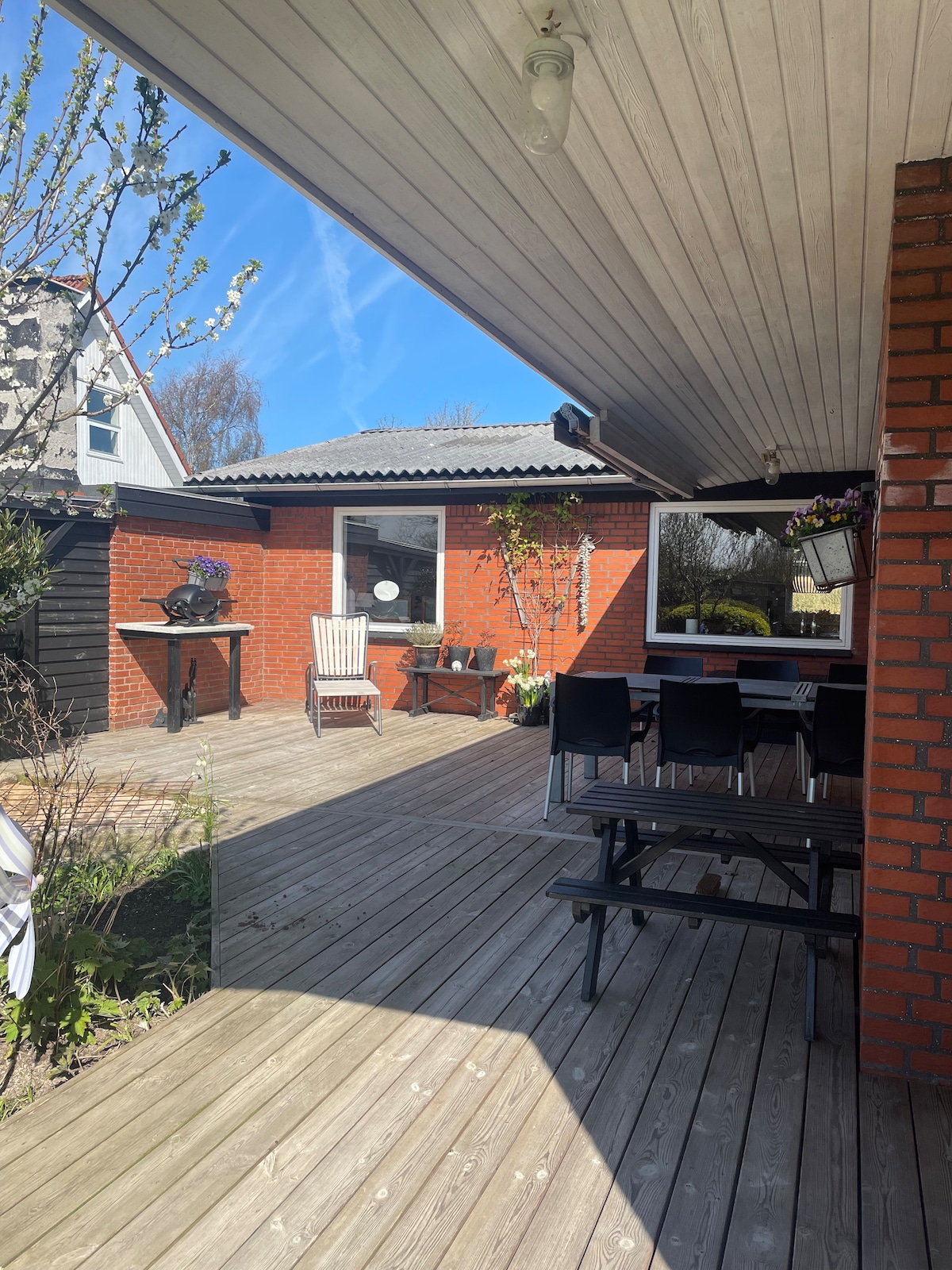 An outdoor patio area features wooden decking and is complemented by brick walls. A dining table with several chairs sits beneath a roofed section, while additional seating is provided by a white rocking chair. Lush greenery is visible along the edges.