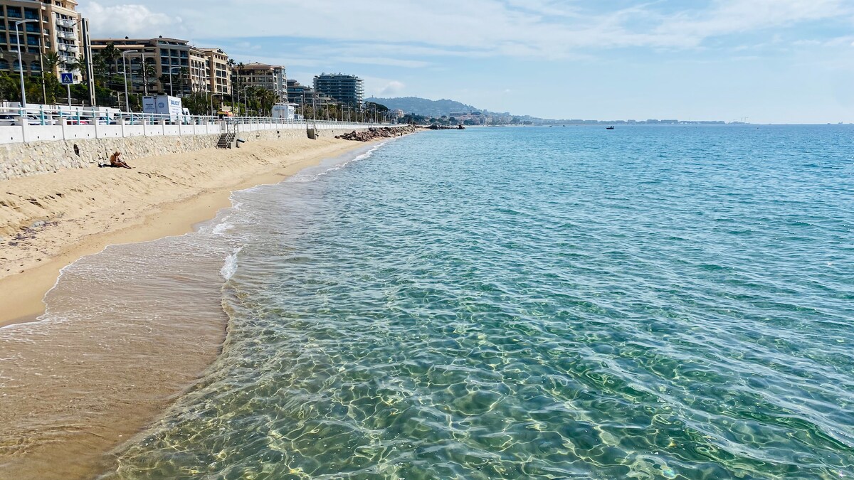 The image captures a serene beach scene featuring a gently sloping sandy shore leading to clear blue water. The coastline is lined with distant tall buildings, with hills visible in the background under a partly cloudy sky.