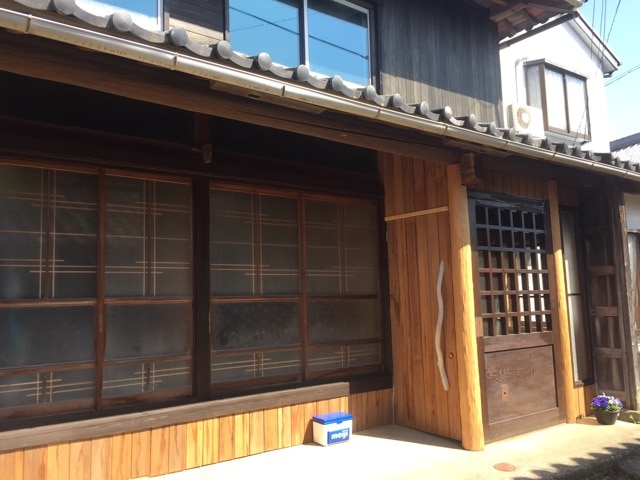 A traditional wooden building features a charming facade with multiple large windows adorned with simple grid patterns. The entrance is highlighted by a wooden door with a grid design, complemented by a wooden bench beneath the windows. Natural light filters through the windows, creating a warm ambiance.