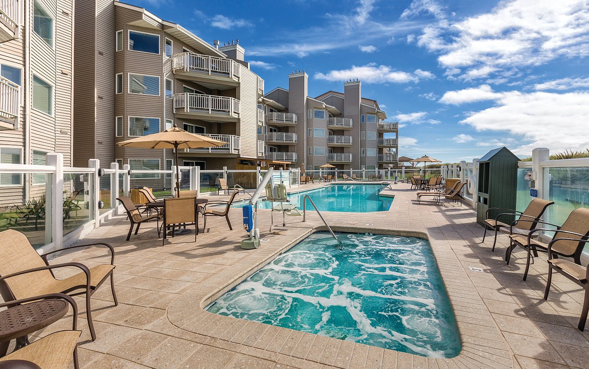 An outdoor pool area is surrounded by multiple lounge chairs and umbrellas. A hot tub is positioned beside the pool, with bubbles visible in the water. The backdrop includes two multi-story buildings with balconies under a clear blue sky.